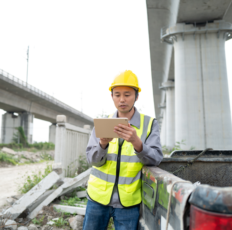 A construction worker wearing a hard hat and safety vest uses a tablet computer.