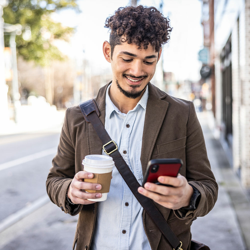 Young man walking down the street holding a coffee and smiling while looking at his phone 