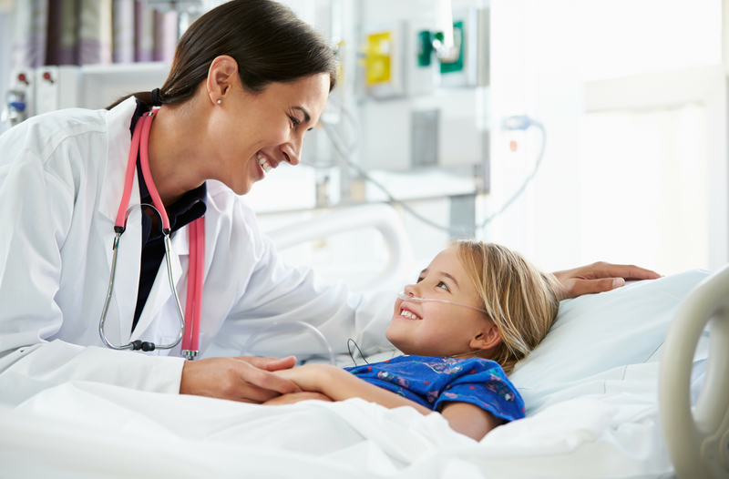  A smiling doctor wearing a stethoscope leans over a young child lying in a hospital bed.