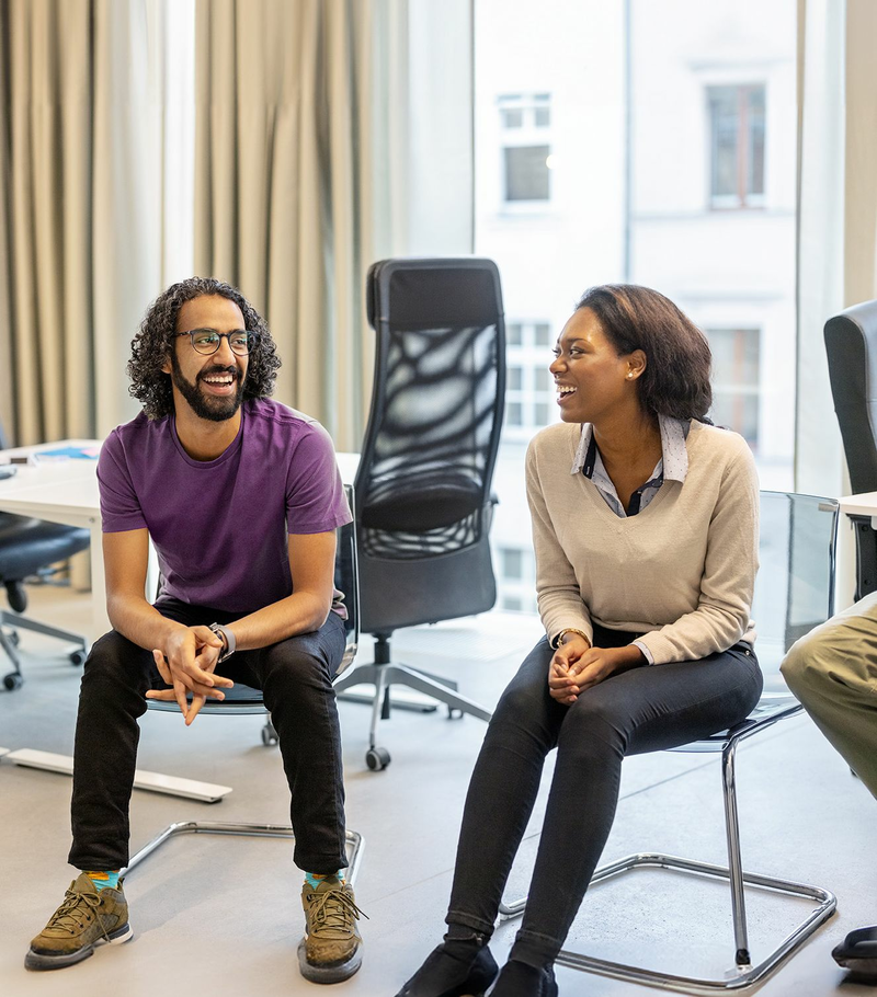 Two people are sitting on chairs in an office, looking at each other, and smiling.