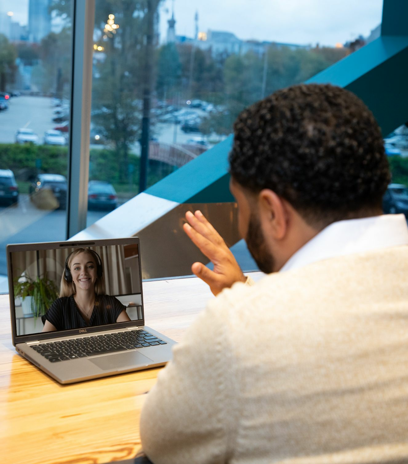 A man is sitting with his back to the camera, having a video call with a coworker on his laptop.