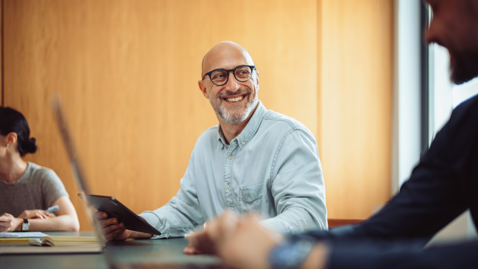 A man with glasses holding a tablet in an office setting and smiling at the person next to him