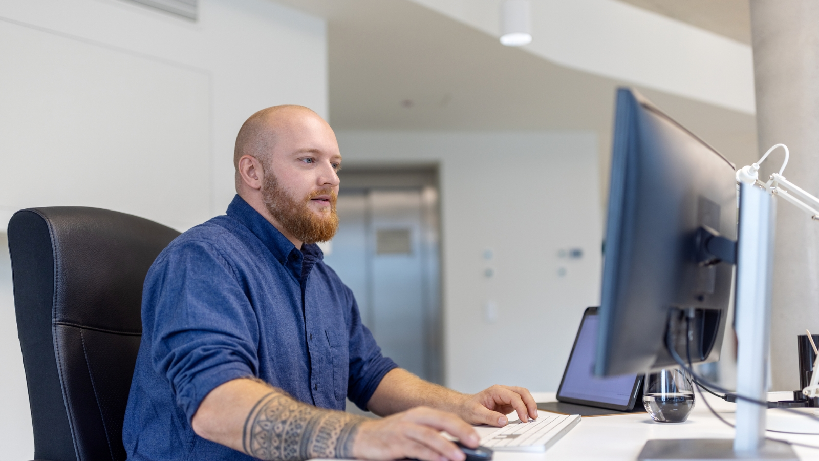 Bearded man in blue shirt works at his desk using a computer and keyboard.