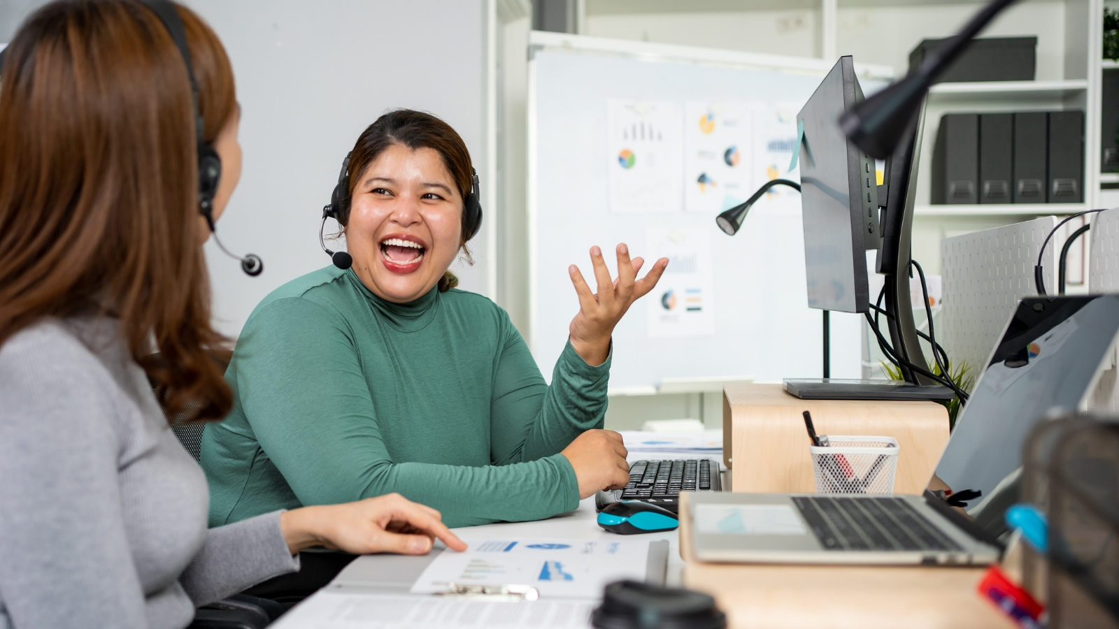 Two businesswomen wearing headsets work together at their office desks.