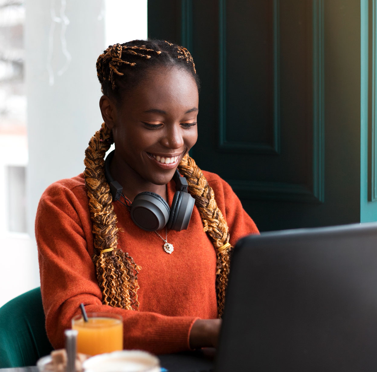  A person with long box braids smiles while using her laptop in a coffee shop.