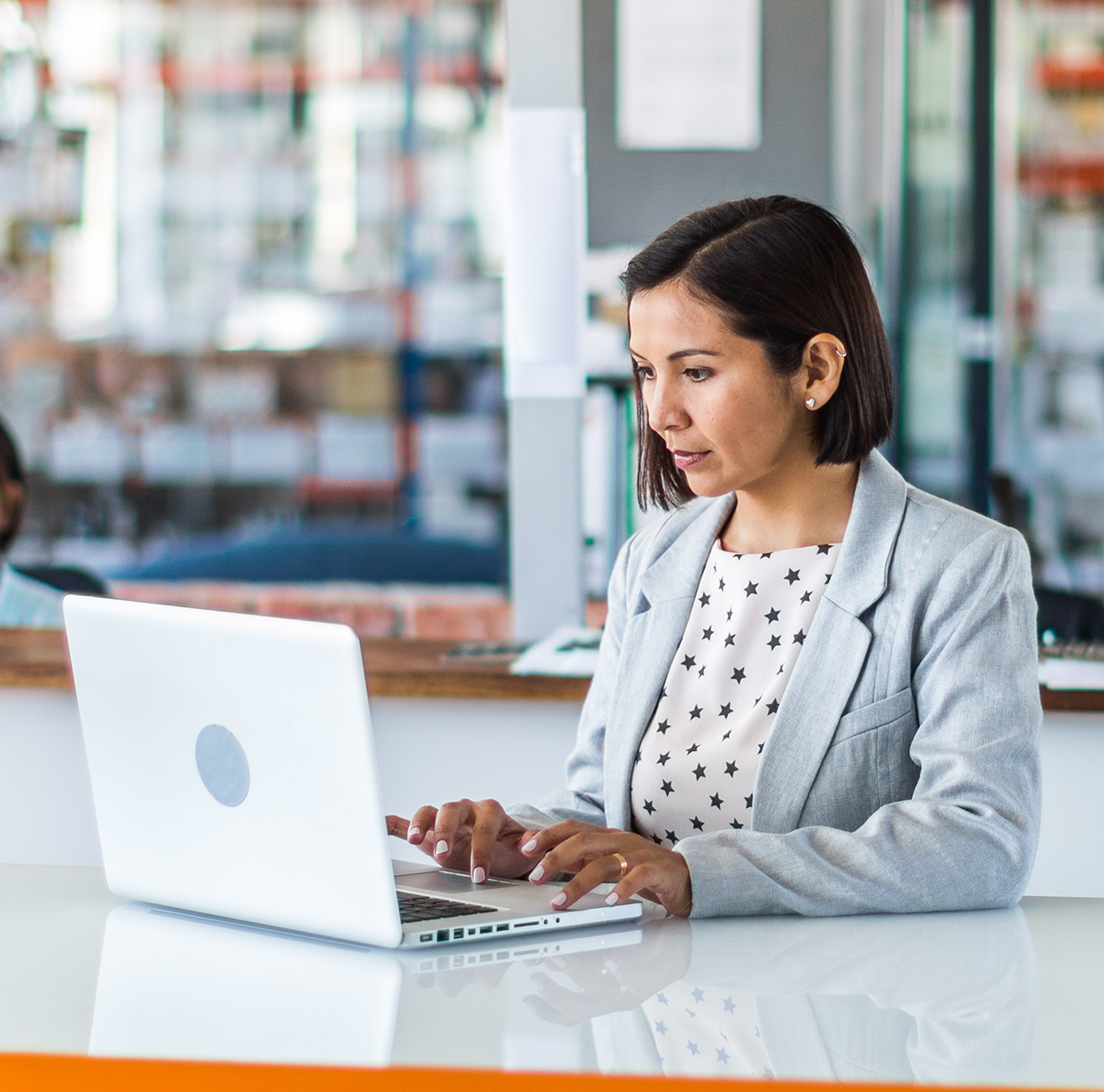 A young woman in a suit is sitting at a desk and working on her laptop.