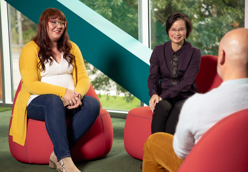 Three people are sitting in chairs in a brightly lit room, looking at each other, and talking.