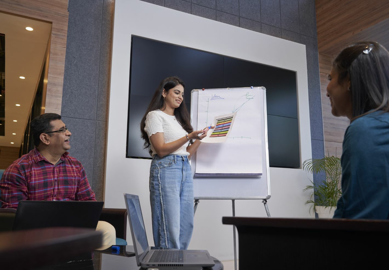 A young woman stands in front of a flipchart giving a presentation to two colleagues.