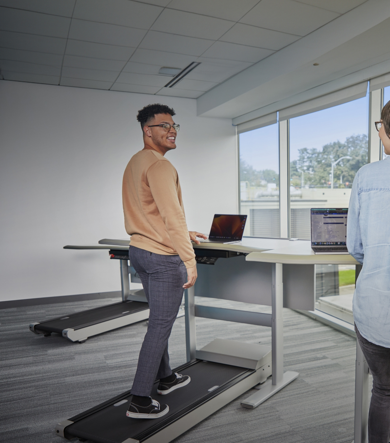 A man in business casual clothing is walking on a treadmill desk while looking at his laptop.