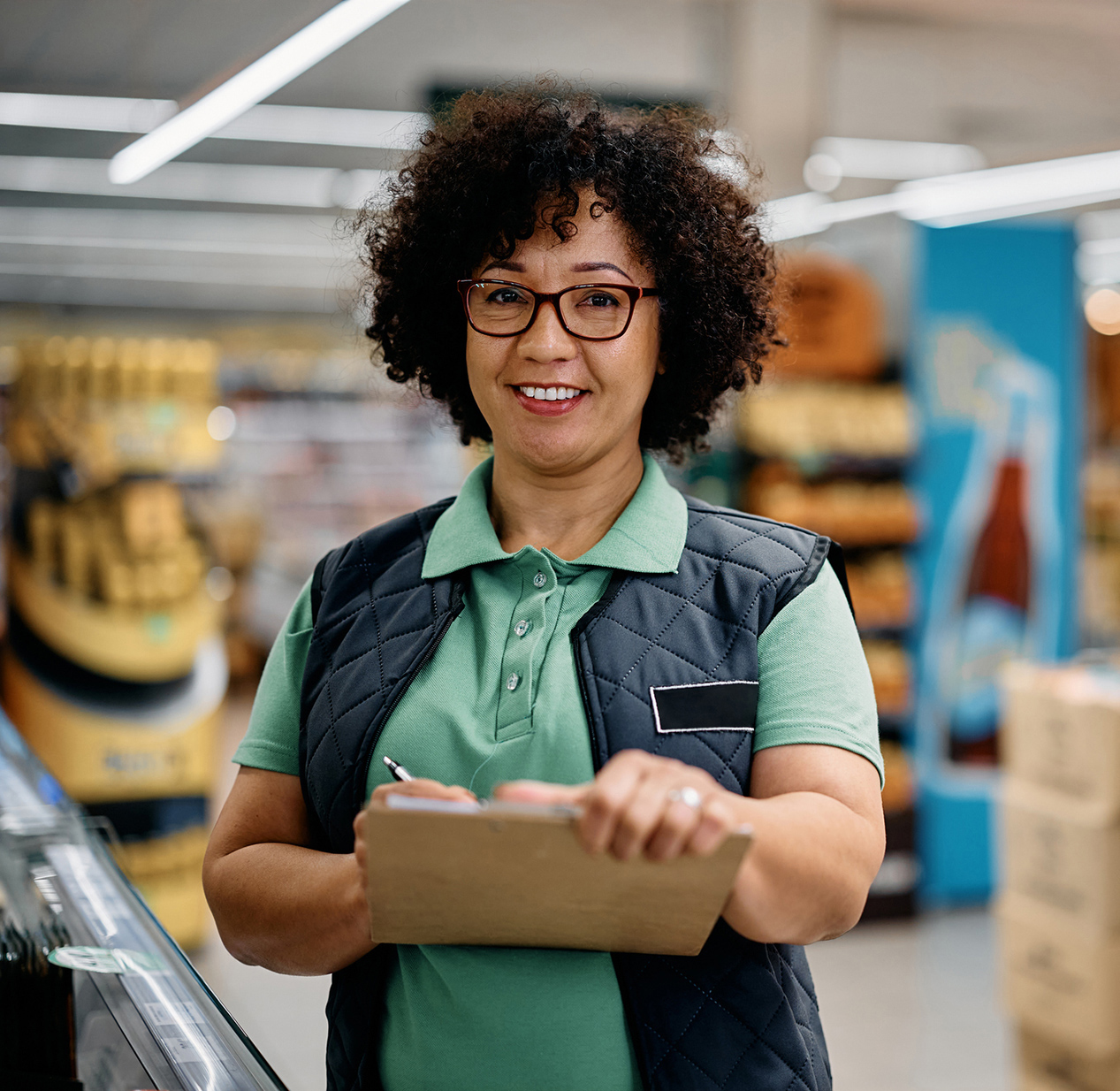 A smiling woman wearing glasses is standing in a hardware store, writing on clipboard