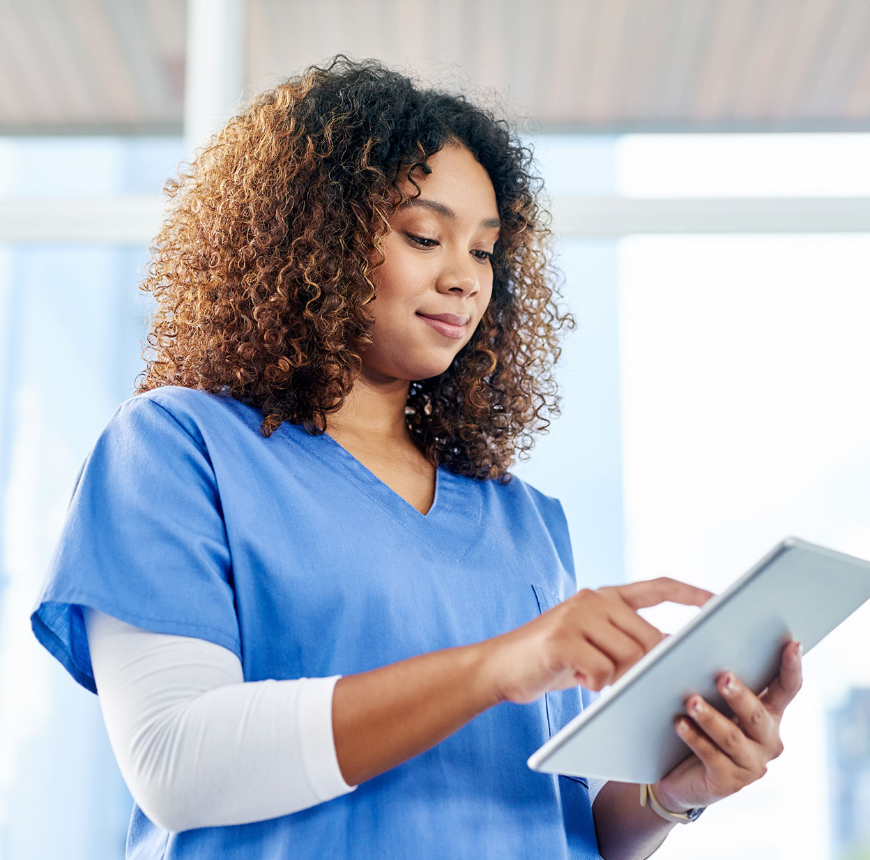 A young female nurse wearing blue scrubs is using a tablet.