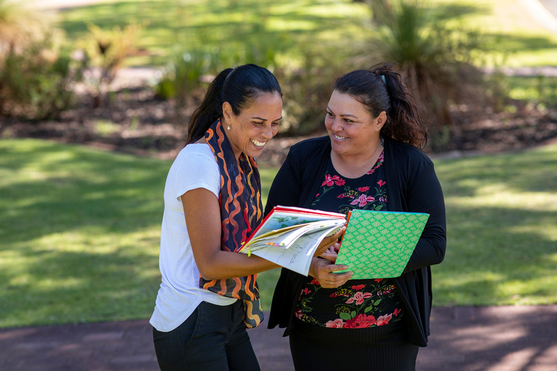  Two smiling students, one holding a book and the other holding a notebook, are standing together