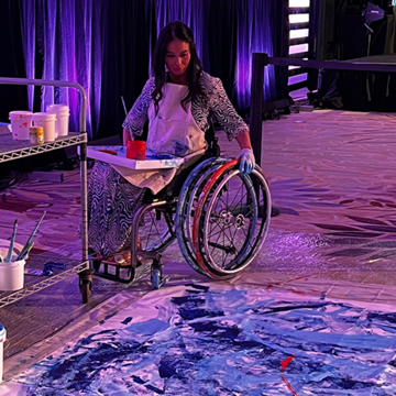  A woman in a wheelchair uses her hand to paint on a large canvas on the floor.