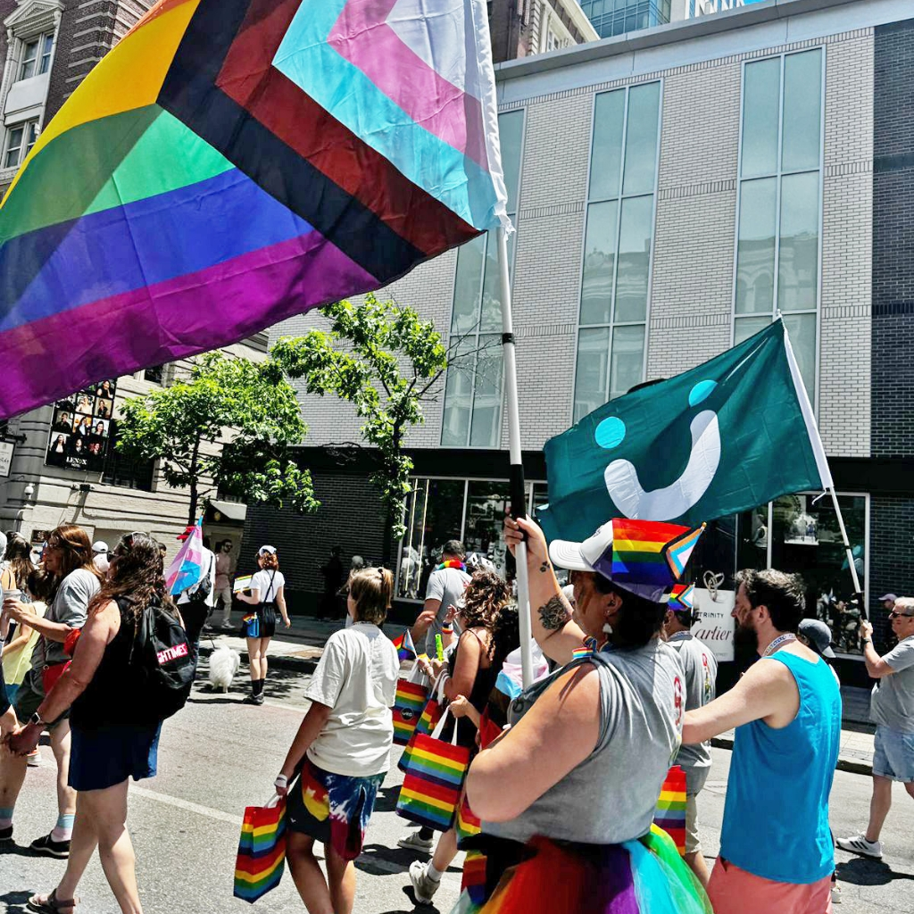 Pride flags and corporate logos wave in the air during a parade.