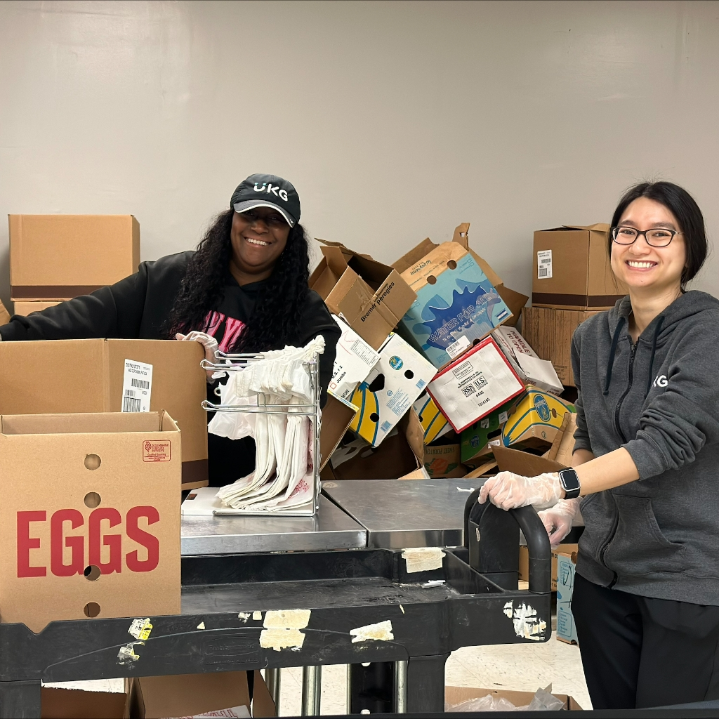 Two smiling women wearing gloves stand behind a table full of boxes and a rack of grocery bags.