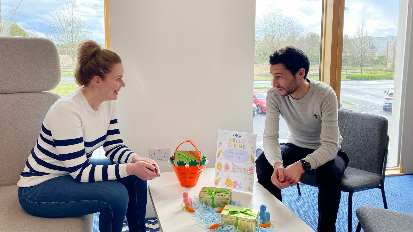 Two people are sitting on a sofa in an office space, looking at each other and smiling.