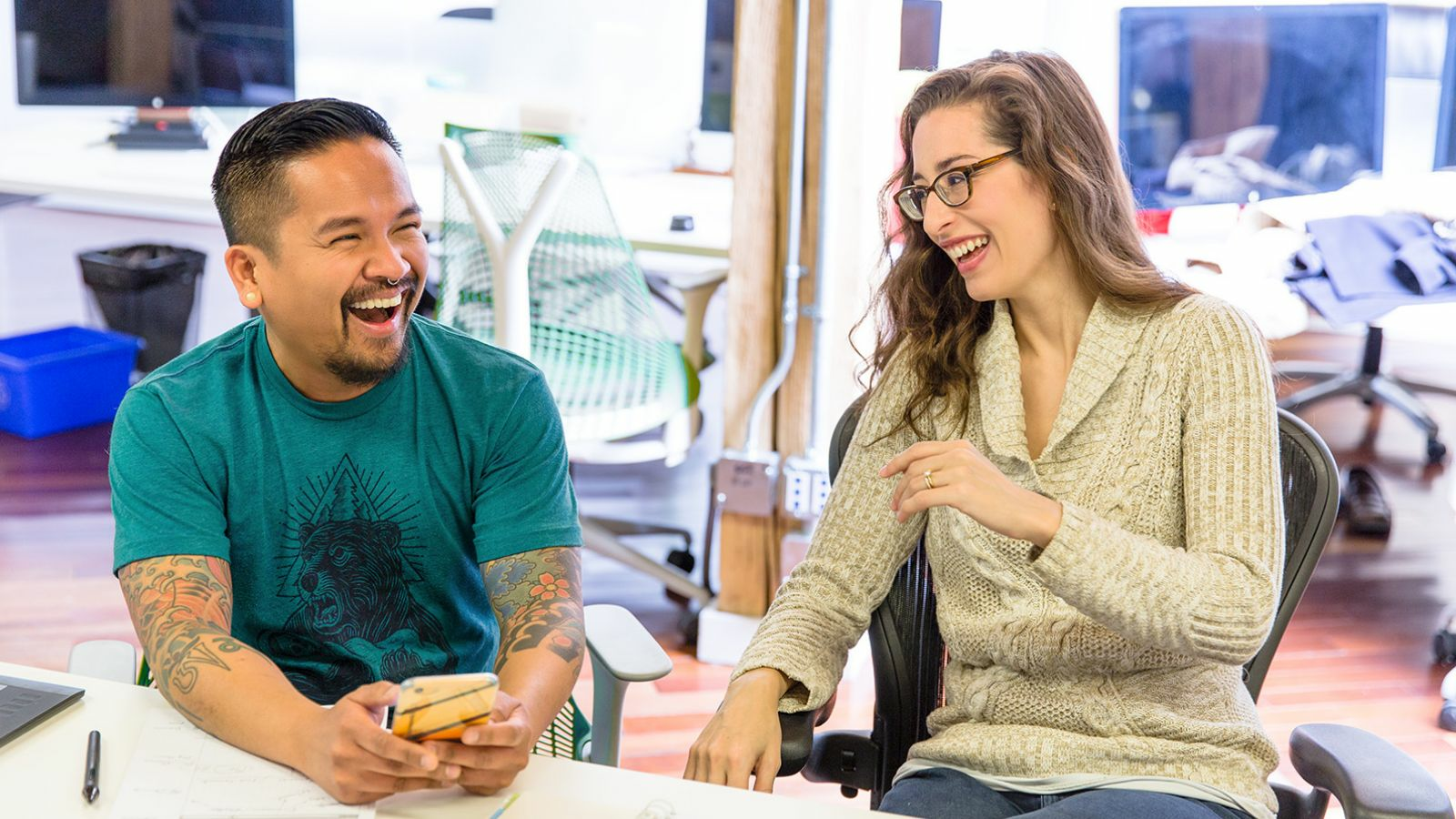 Two coworkers are sitting in chairs and laughing at something on a phone.