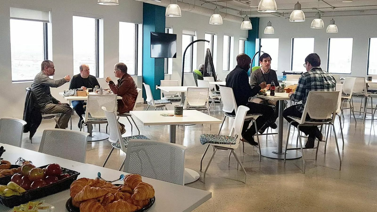 A group of six men are eating lunch and talking in a modern office break room.