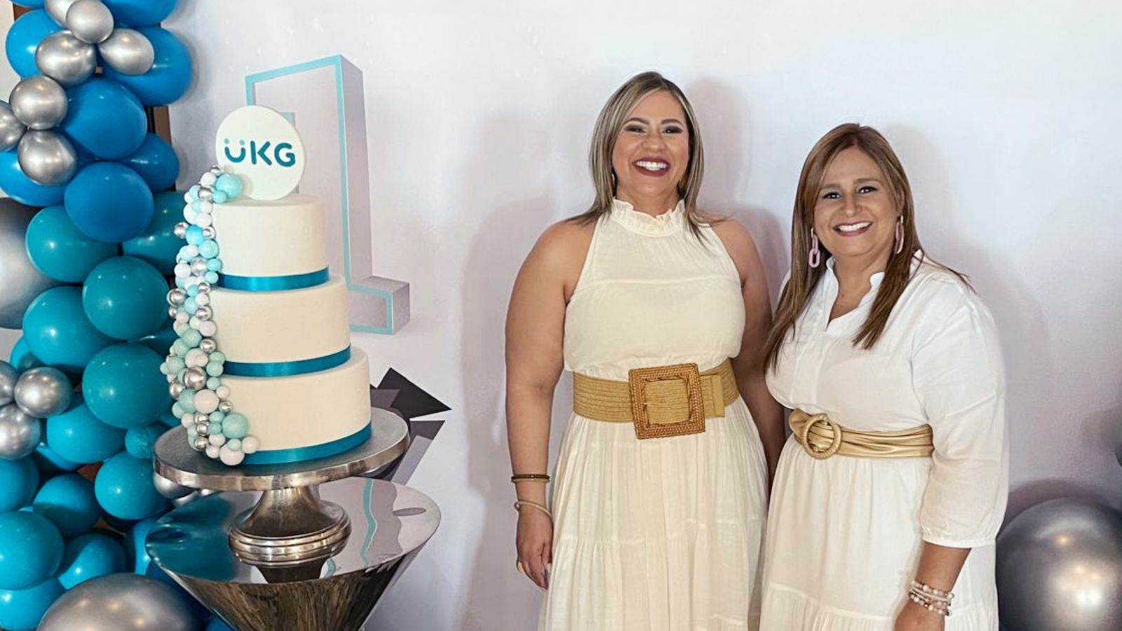 Two smiling women in white dresses stand in front of a UKG tiered cake.