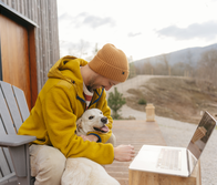 A man wearing a yellow jacket and beanie sits on a porch with his dog and uses a laptop.