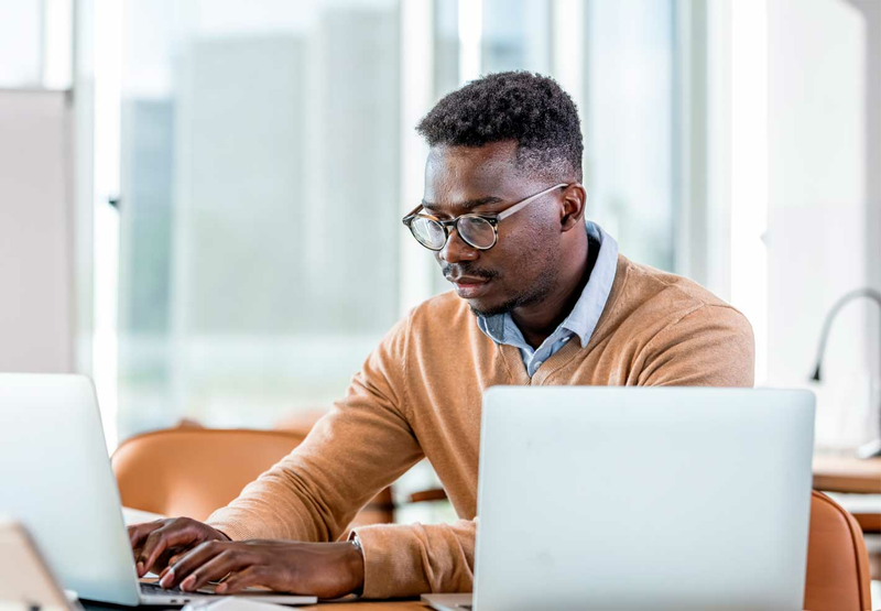 A young man wearing glasses sits at a desk and looks at two laptops.