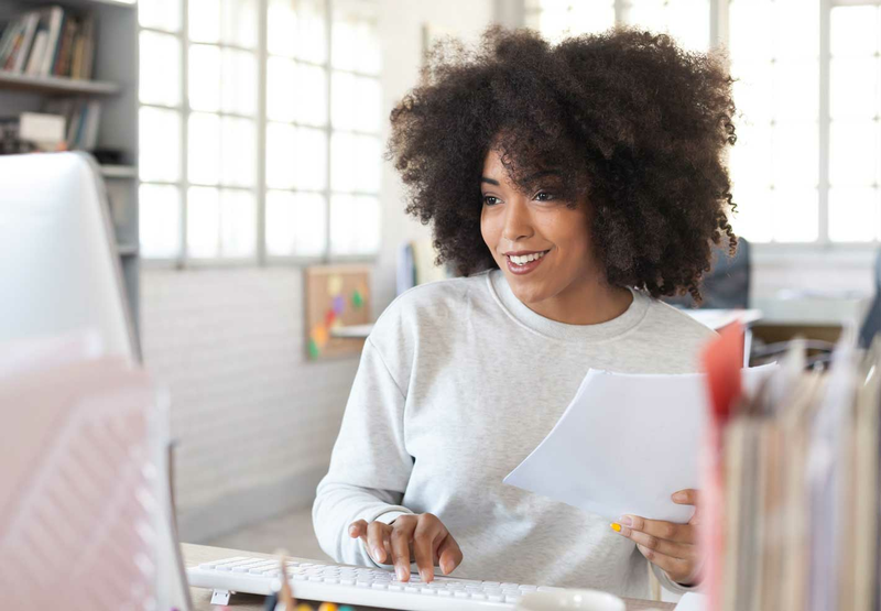 A young woman with curly hair is sitting at her desk looking at a computer monitor