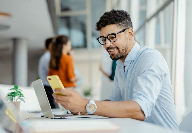 A young professional wearing glasses is sitting at his desk looking at his phone.