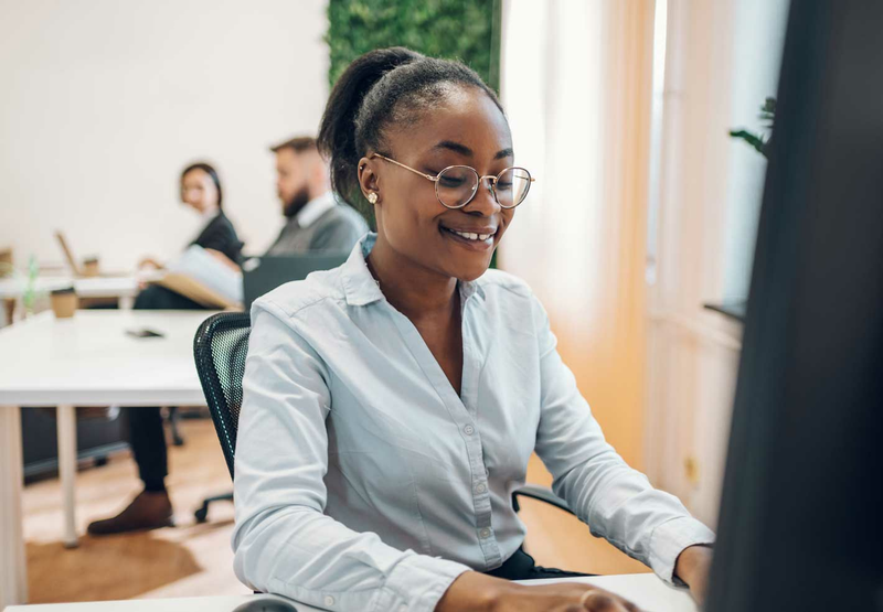 A smiling woman wearing glasses sits at her desk and looks at her computer screen.