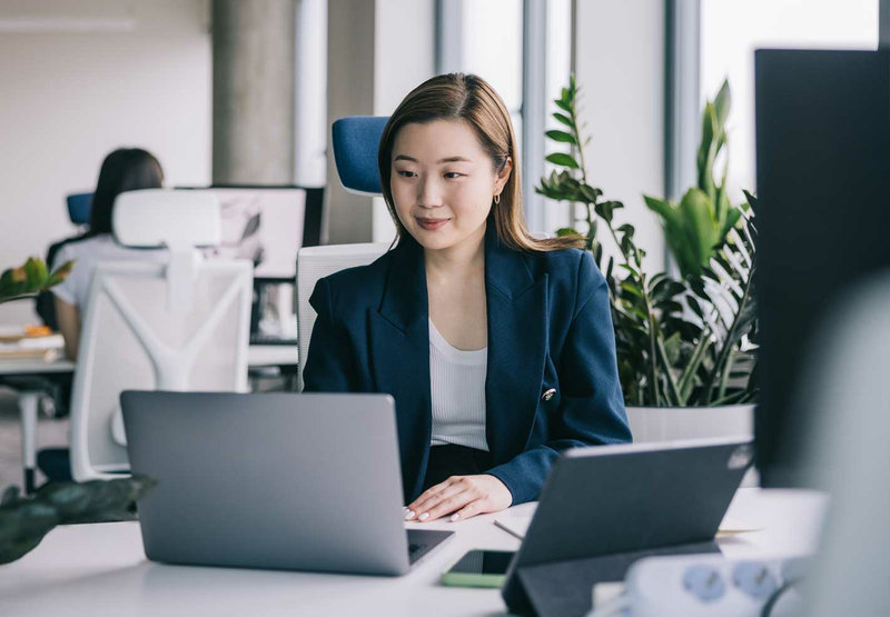 A young woman in a blue suit jacket is sitting at her desk looking at her laptop.