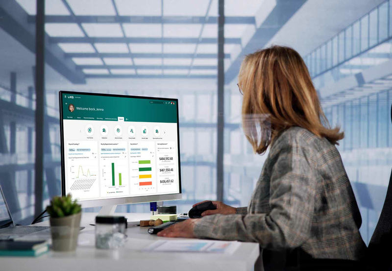 A woman wearing a green jacket is sitting at her desk looking at a computer screen displaying a UKG product.