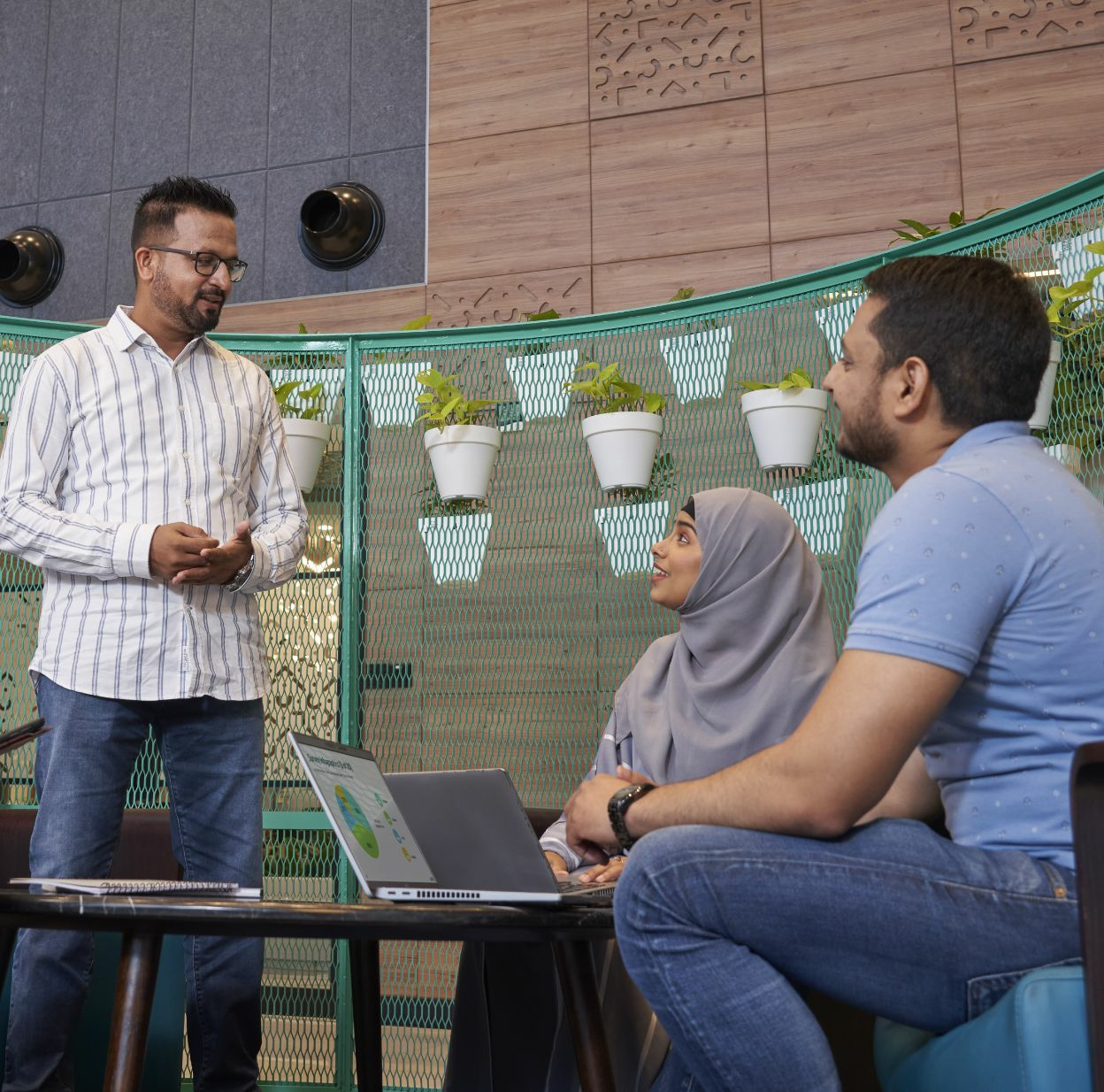 A man stands talking to a man and woman who are sitting at a table.