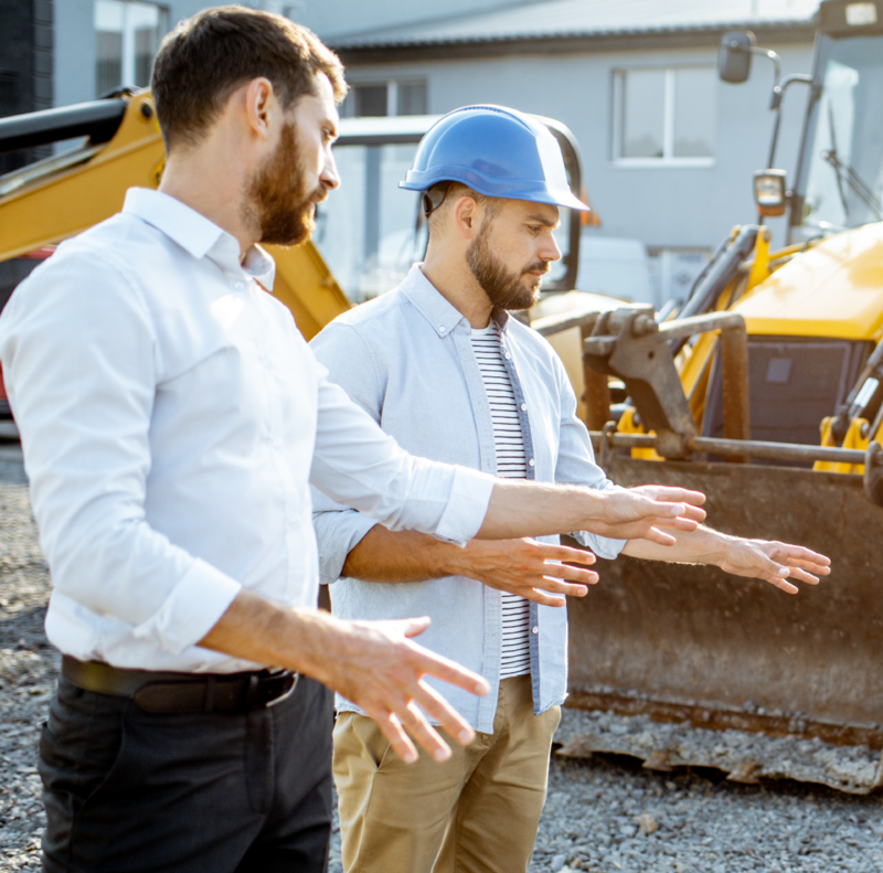 Two men in hard hats are standing next to a backhoe.