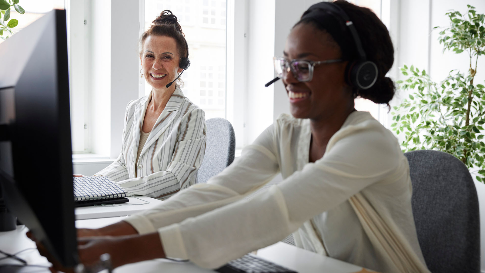 Two women wearing headsets sit at their desks and smile at each other.