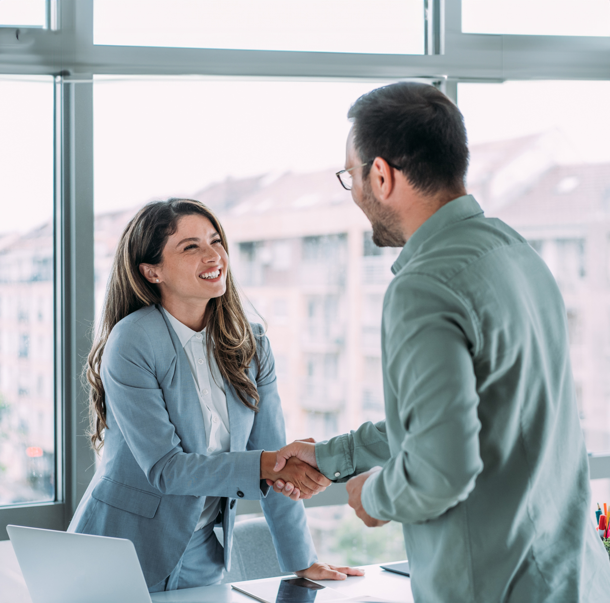  Two business people are standing in an office space, smiling and shaking hands.