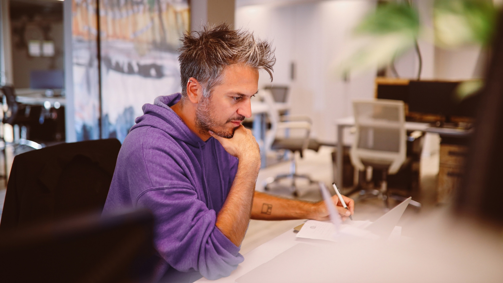 A man wearing a purple hoodie is sitting at a desk in an office, looking at a laptop.