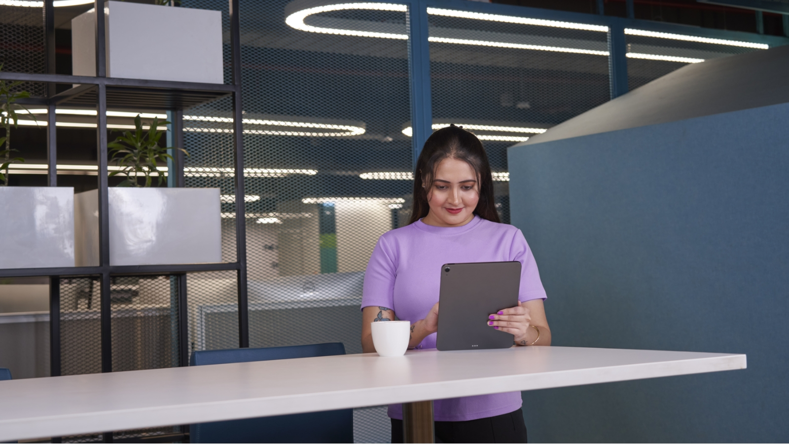 A young woman in a purple shirt is standing in an office, looking at a tablet.