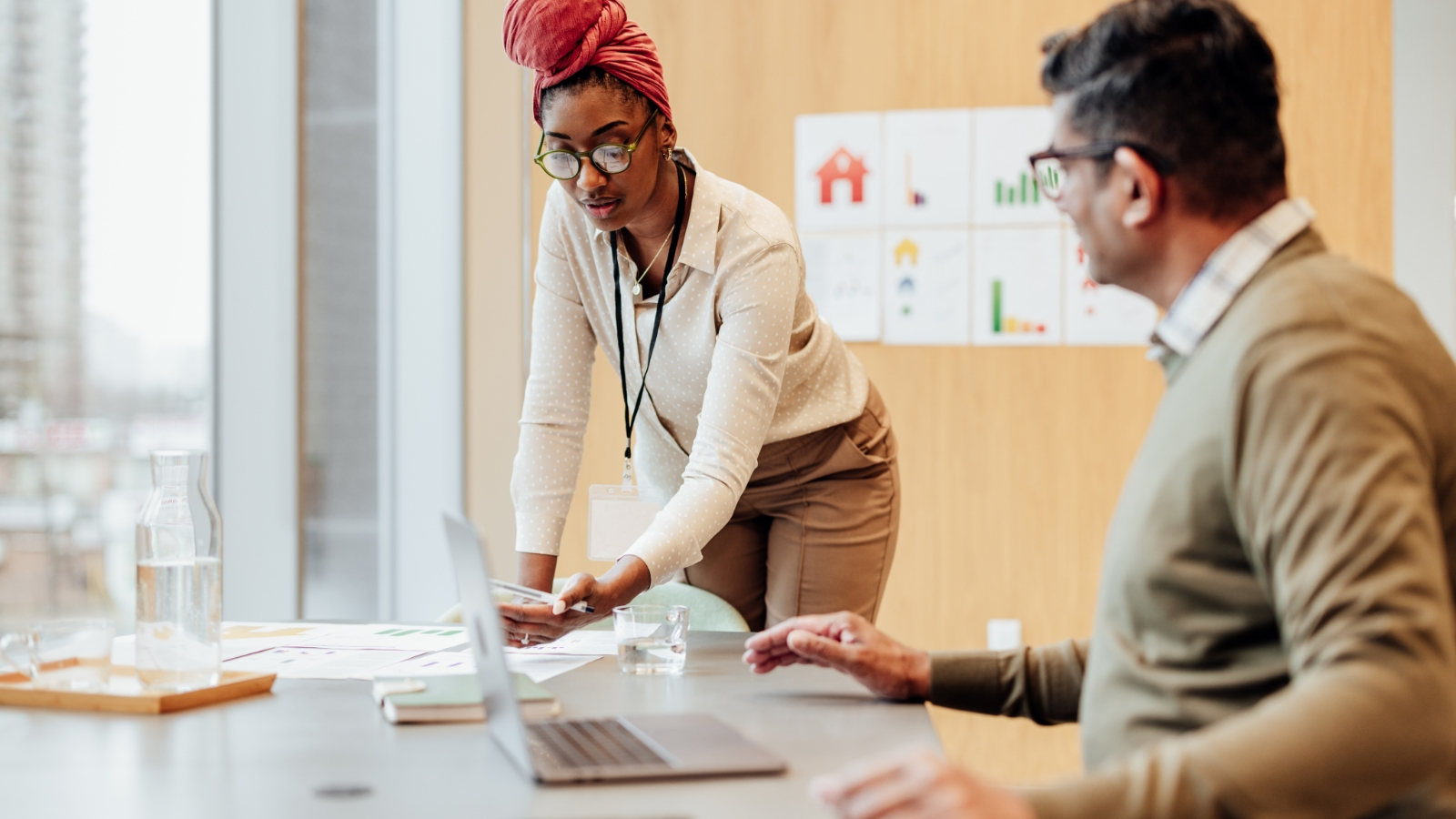  A woman in a pink head wrap and glasses is showing a man in a brown sweater something on her laptop.