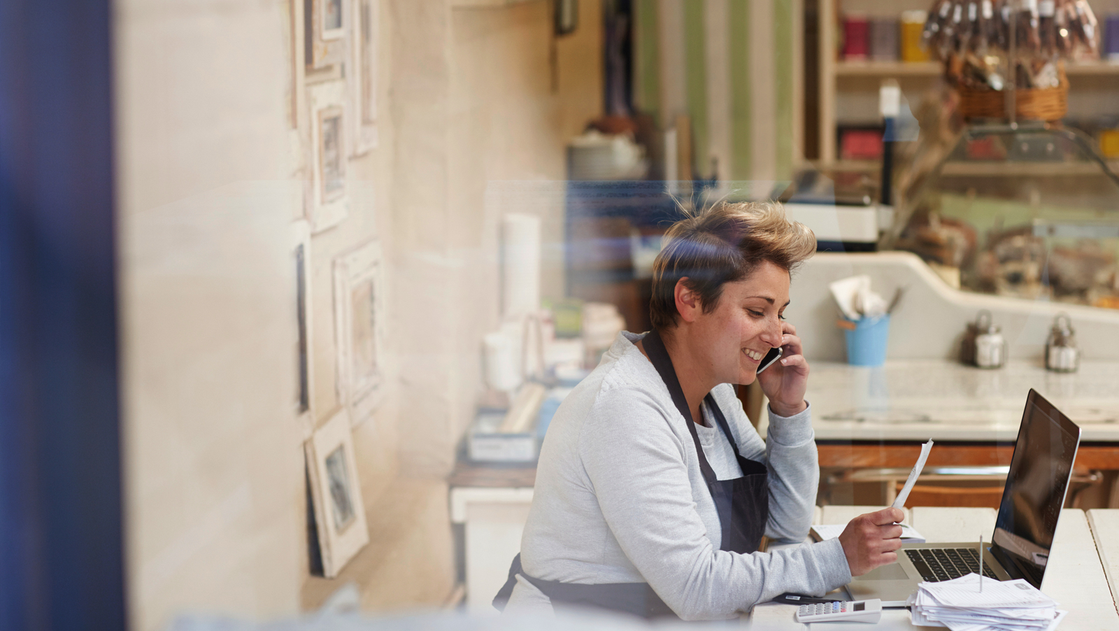  A woman wearing an apron is sitting at a table in a cafe, looking at a laptop and smiling